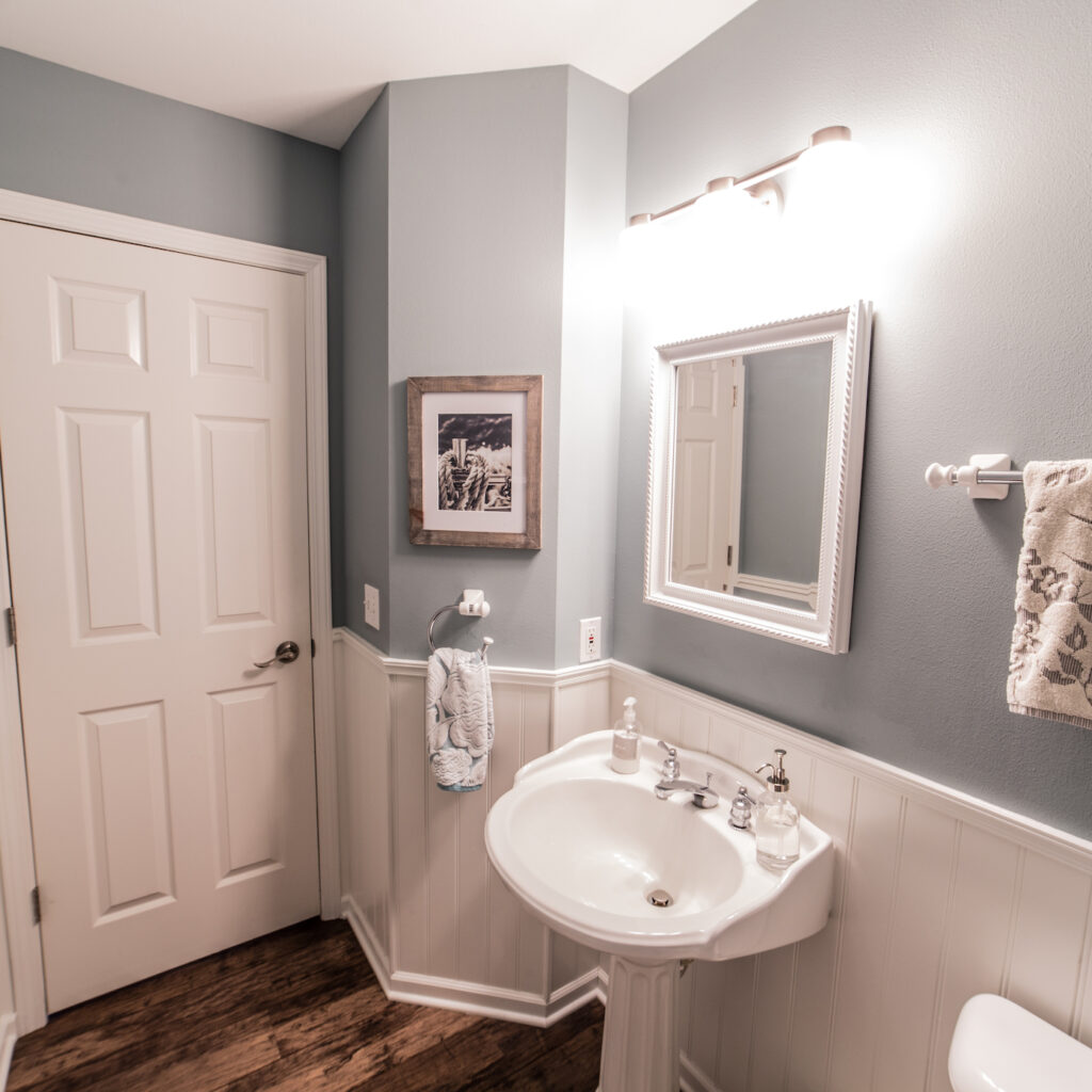 Remodeled half bathroom with porcelain pedestal sink, white framed mirror, nickel plated light fixture and white wooden panel wainscoting.