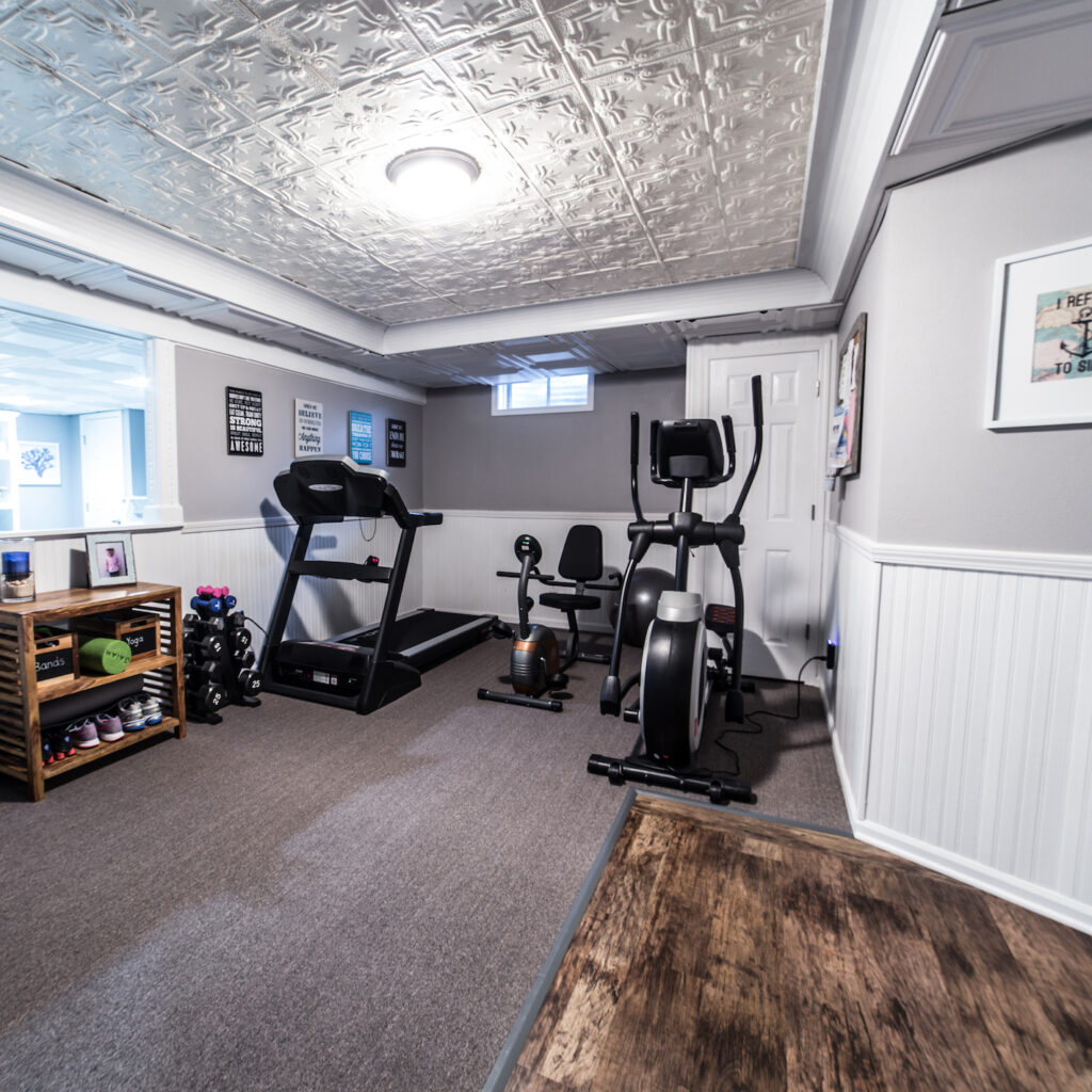 Half basement remodeled and converted into an exercise room with white wooden paneled wainscoting, Faux tin ceilings, partially carpeted flooring, partially laminated wood flooring, and exercise equipment.