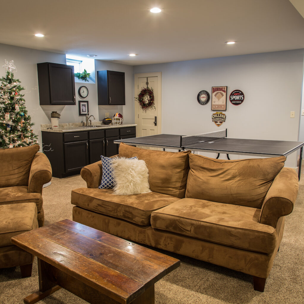 Home remodel with basement converted into living space consisting of wet bar, black cabinets, ping pong table, brown suede couch and chaise lounge chair.