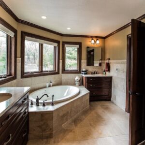 Twin cherry vanities with light grey granite countertops and bronze faucet fixtures separated by a fully tiled jetted porcelain bathtub in a fully remodeled master suite bathroom.
